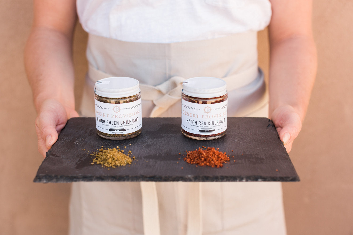 Person holding a tray containing a jar of hatch green chile salt and hatch red chile salt and piles of each to show texture